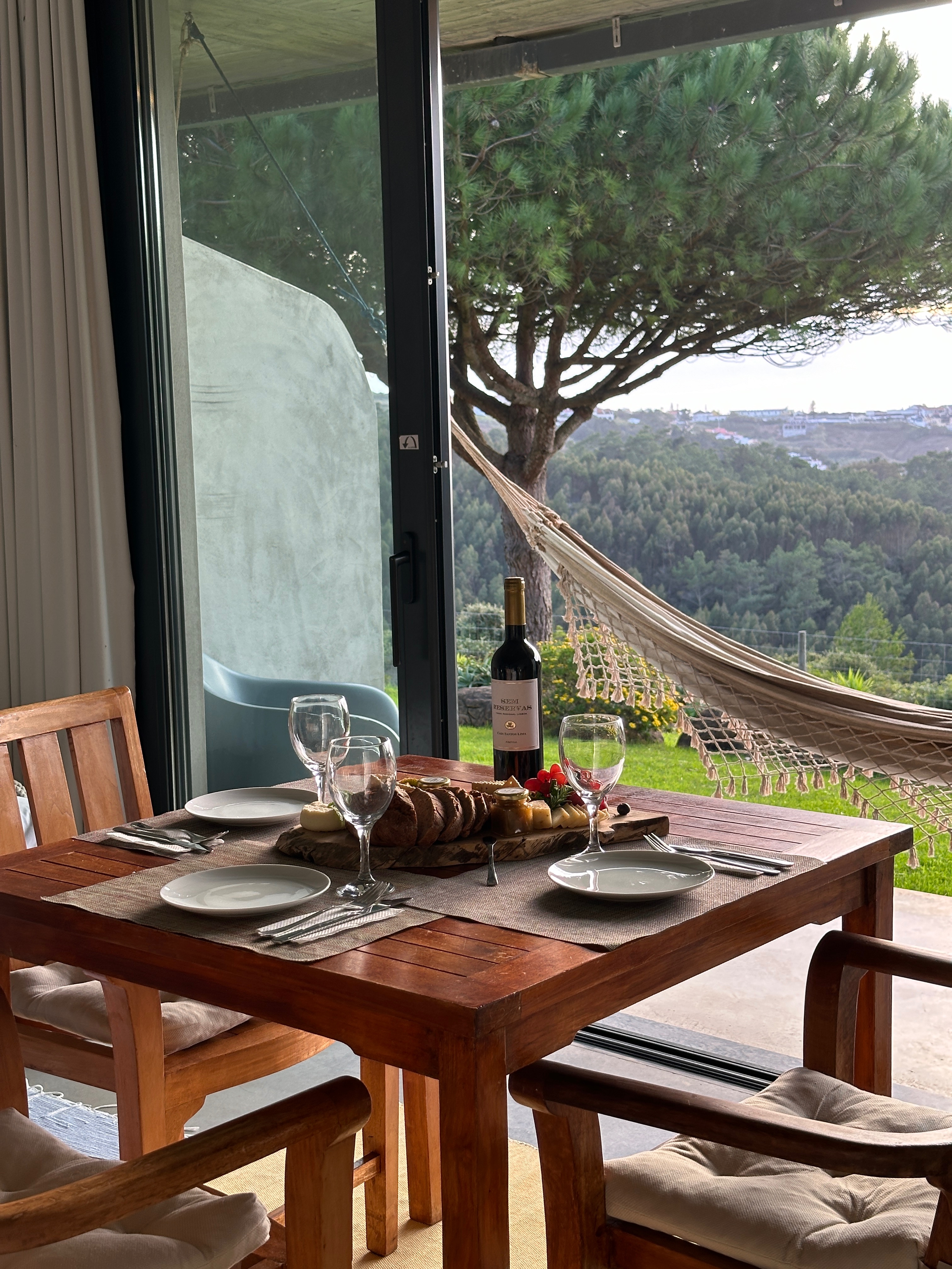 Intimate dining table set for two with wine glasses, overlooking a garden with hammock through glass doors