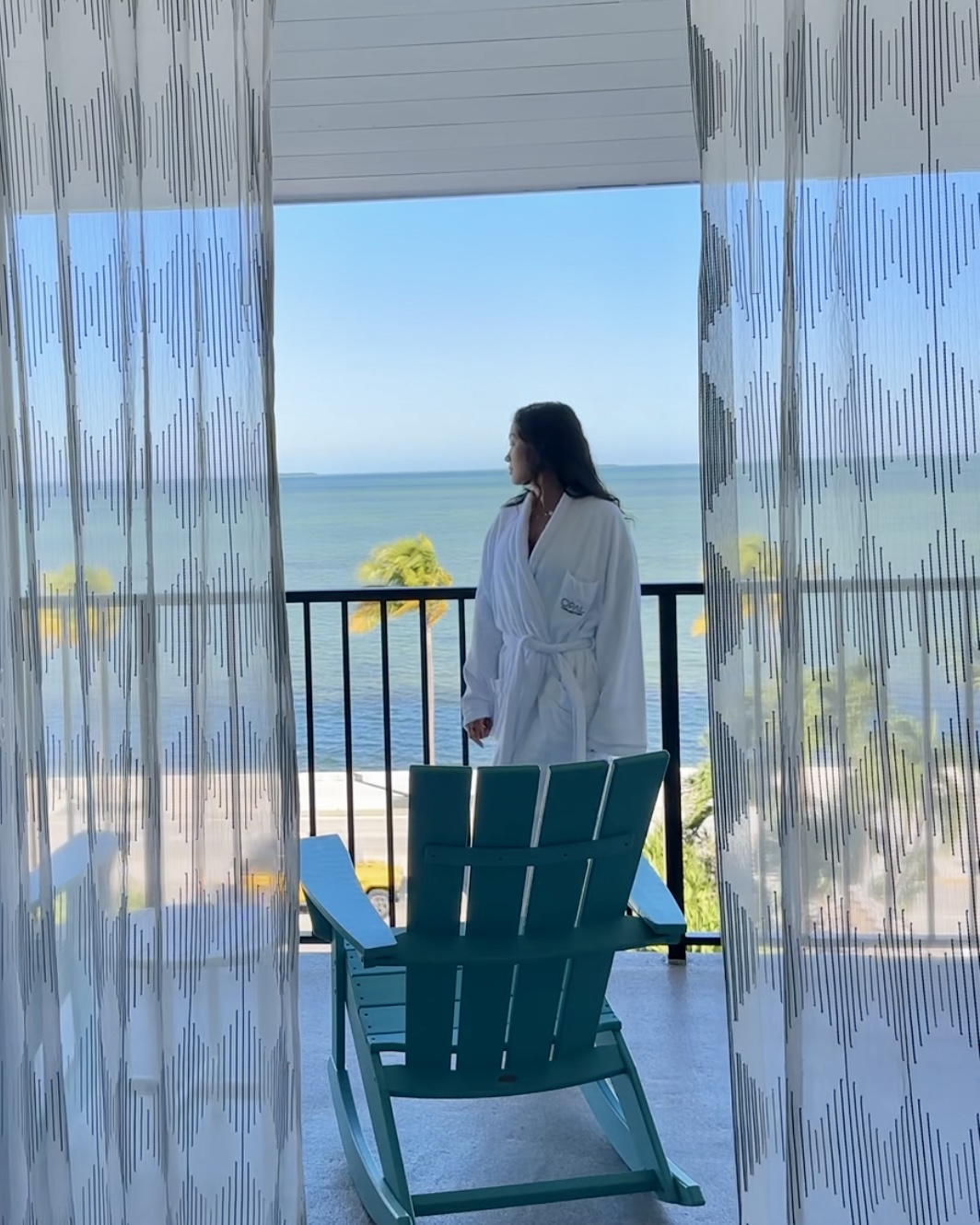 Nianch in a white hotel robe on a balcony overlooking the sea, patterned curtains and a teal Adirondack chair in the foreground